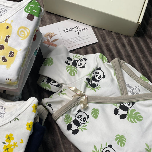 A baby clothing set laid out on a dark ribbed fabric background, including a white zip-front romper with black-and-white panda prints and green leaves, folded white and yellow patterned fabric pieces, a cream-colored box with the brand name 'monda babu', and a small plush toy.